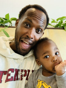 Njuguna and his toddler pose together. Njuguna is smiling. The baby has a thumb in his mouth.