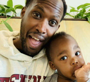 Njuguna and his toddler pose together. Njuguna is smiling. The baby has a thumb in his mouth.