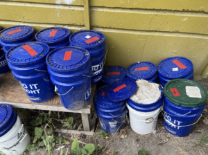 Blue buckets on the ground and on a bench with labels on their lids.