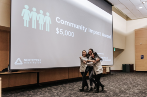 Three Asian American women hug each other in front of a large screen with the words "Community Impact Award $5,000" projected onto it.