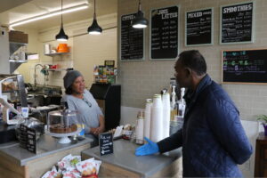 A Black man wearing latex gloves speaks to a Black woman standing behind a cafe counter.