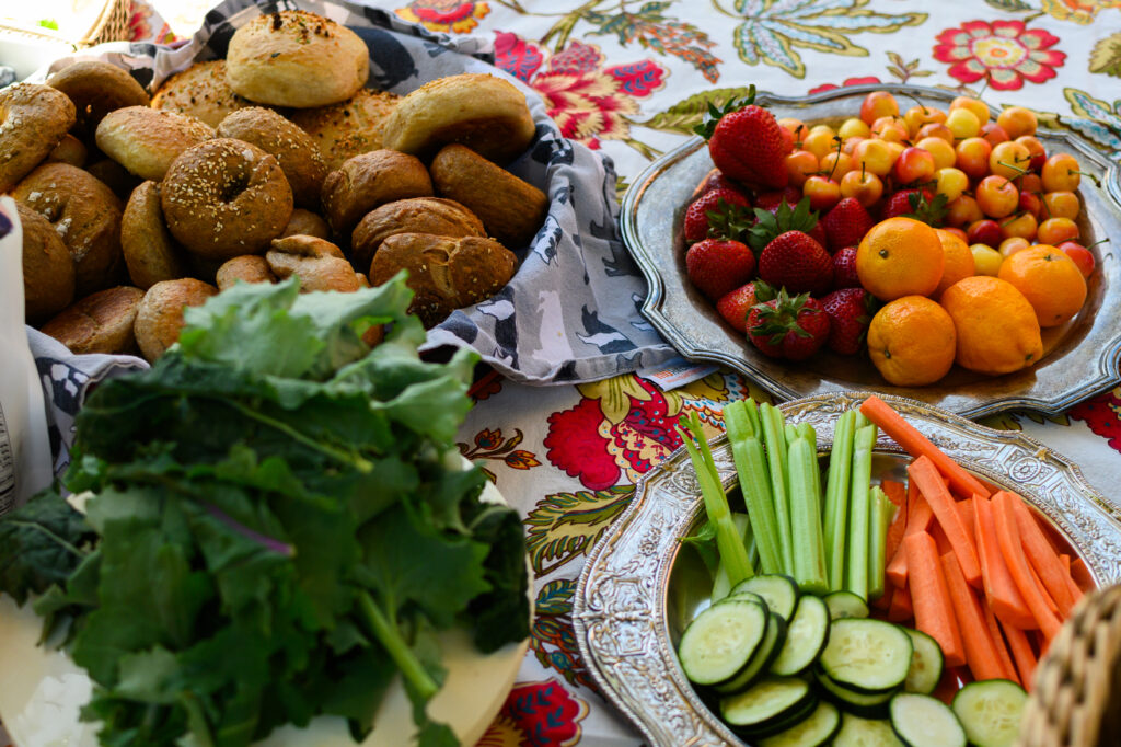 Platters with homemade bagels, fruit, and vegetables on a floral tablecloth.